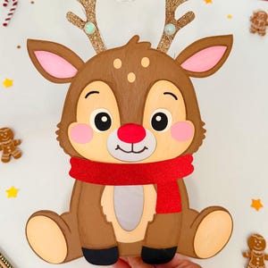 May include: A brown reindeer decoration with a red scarf, glittery gold antlers, and a red nose. The reindeer is surrounded by gingerbread men and star-shaped decorations, perfect for Christmas.