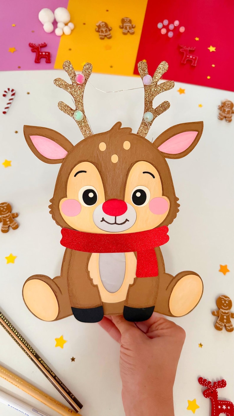 May include: A brown reindeer decoration with a red scarf, glittery gold antlers, and a red nose. The reindeer is surrounded by gingerbread men and star-shaped decorations, perfect for Christmas.
