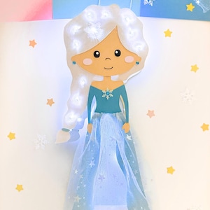 Könnte beinhalten: Eine hellblaue Prinzessinnen-Bastelarbeit mit einem weißen Tüllrock und einem weißen Zopf mit weißen Lichtern. Die Prinzessin hat ein hellbraunes Gesicht mit einem hellrosa Rouge und ein hellblaues Kleid mit einem Schneeflockenmuster. Der Hintergrund ist weiß mit gelben Sternen.