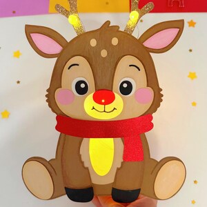 May include: A brown reindeer-shaped Christmas decoration with a red scarf and a glowing red nose. The reindeer has glittery gold antlers with small lights. The decoration is held by a hand and surrounded by festive elements like gingerbread men and stars.