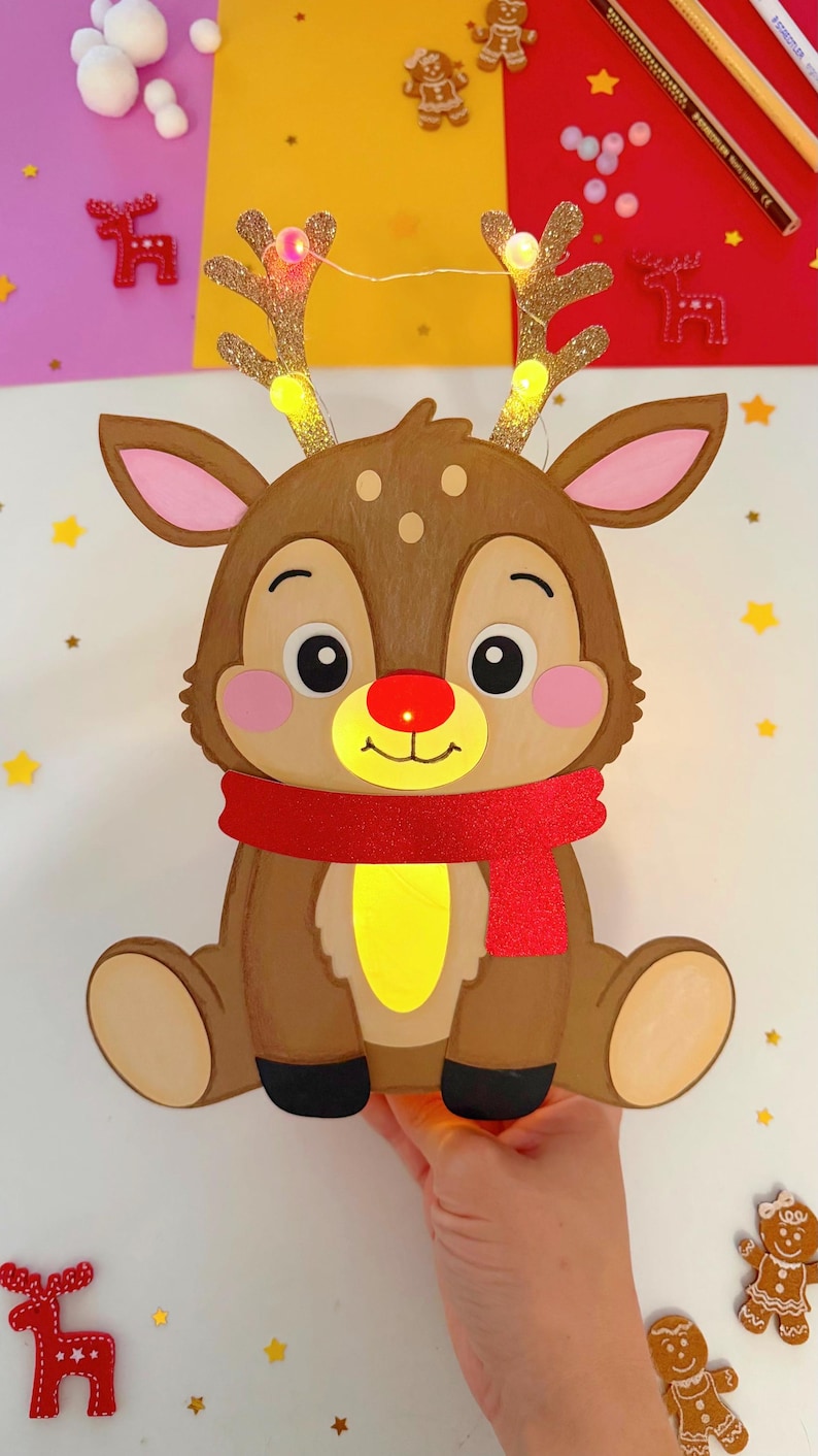 May include: A brown reindeer-shaped Christmas decoration with a red scarf and a glowing red nose. The reindeer has glittery gold antlers with small lights. The decoration is held by a hand and surrounded by festive elements like gingerbread men and stars.