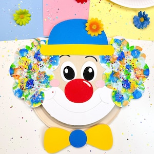 May include: A paper plate craft of a clown with a blue hat, yellow bow tie, and a red nose. The clown's face is painted with colorful splatter paint.