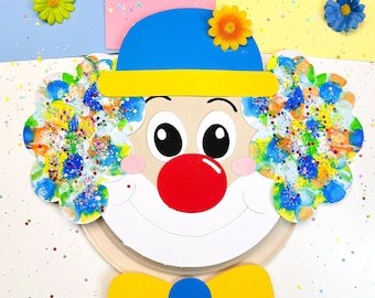 May include: A paper plate craft of a clown with a blue hat, yellow bow tie, and a red nose. The clown's face is painted with colorful splatter paint.