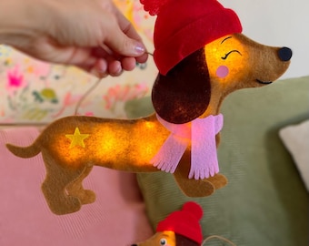 May include: Two felt dachshund ornaments with internal lights. The larger one wears a red pom-pom hat and a pink scarf, while the smaller one wears a red hat and a blue scarf. Both feature a yellow star detail on their brown bodies.