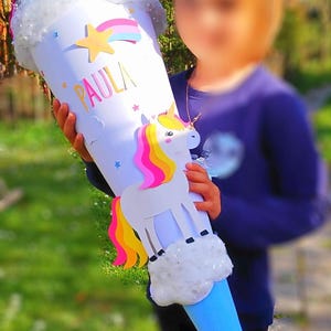 May include: A blue and white unicorn school cone with the name "PAULA" written on it. The cone is decorated with a rainbow, stars, and a unicorn with a pink and yellow mane.