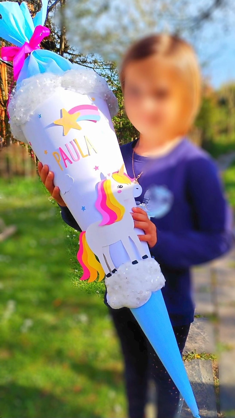 May include: A blue and white unicorn school cone with the name "PAULA" written on it. The cone is decorated with a rainbow, stars, and a unicorn with a pink and yellow mane.