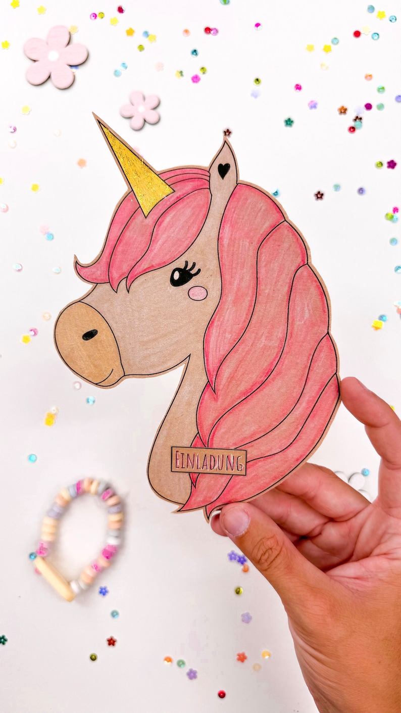 Puede incluir: Un recorte de unicornio rosa y marr&oacute;n con un cuerno dorado y la palabra "Einladung" en una etiqueta marr&oacute;n. El unicornio est&aacute; rodeado de confeti de colores.