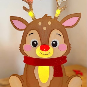 May include: A festive, seated reindeer decoration with a red scarf and glowing yellow belly. The reindeer has a brown body, pink ears, and a red nose. Its antlers are adorned with small, twinkling lights, perfect for holiday decor.
