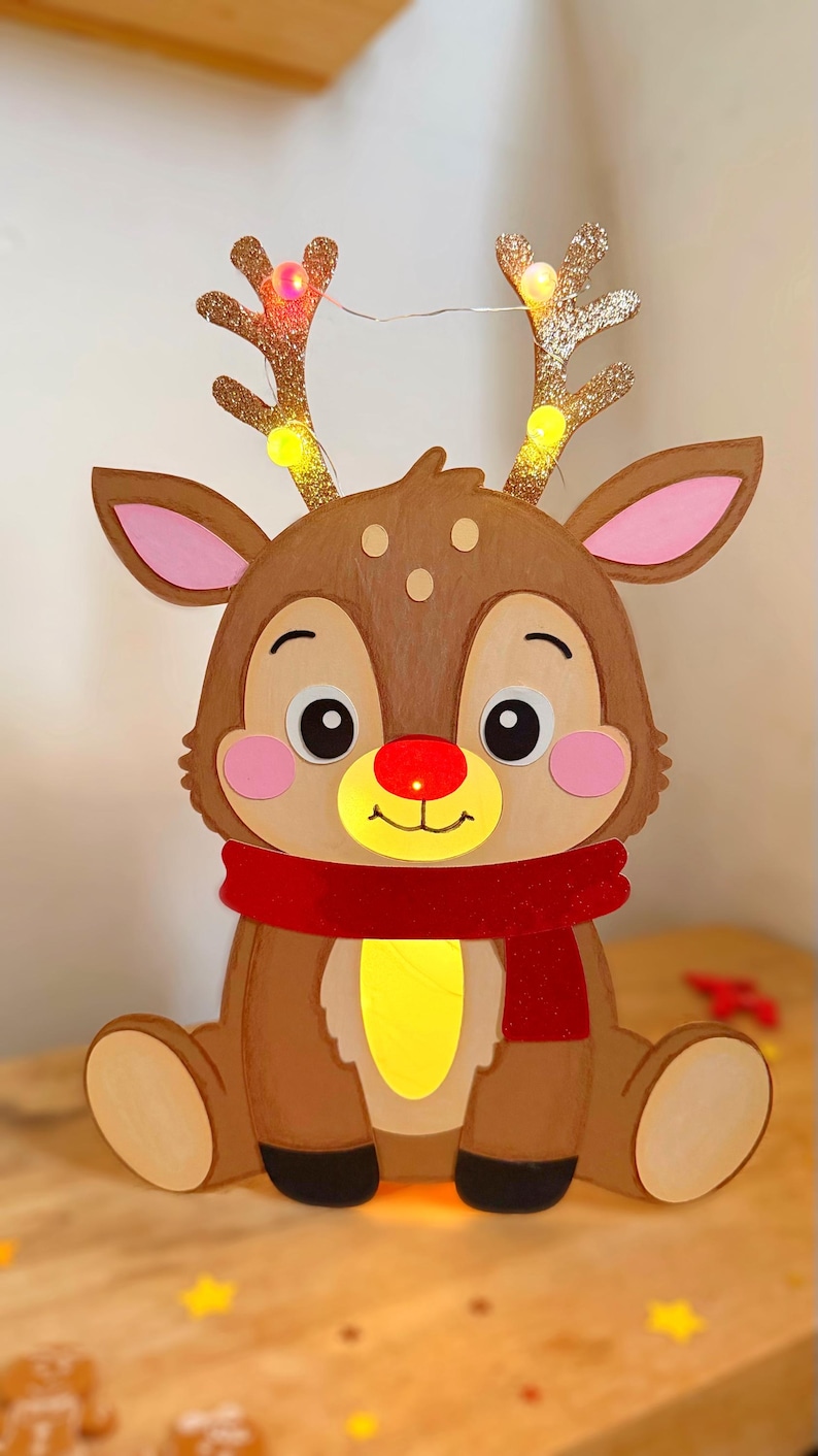 May include: A festive, seated reindeer decoration with a red scarf and glowing yellow belly. The reindeer has a brown body, pink ears, and a red nose. Its antlers are adorned with small, twinkling lights, perfect for holiday decor.