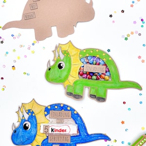Pu&ograve; includere: Tre scatole per feste a forma di dinosauro, una marrone, una verde e una blu. Le scatole sono decorate con dettagli colorati e hanno una finestra ritagliata per contenere leccornie. La scatola verde &egrave; piena di caramelle colorate, la scatola blu ha una barretta di cioccolato Kinder e la scatola marrone ha un testo che dice "Fur", "Wo?" e "Wann?"