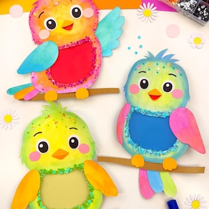 May include: Colorful, handcrafted bird decorations with glitter accents, perched on brown branches. The birds are in shades of yellow, orange, green, and blue. Markers and craft supplies are in the background. A box of crayons is visible.