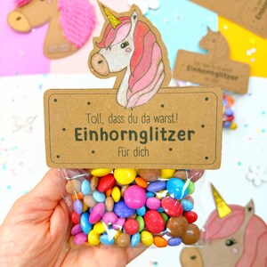 May include: A brown card with a unicorn illustration and the text "Toll, dass du da warst! Einhorn Glitzer f&uuml;r dich."  A clear plastic bag filled with colorful candy is attached to the card.