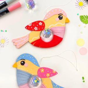 May include: Two handmade bird ornaments. The birds have yellow bodies, red and pink accents, and blue wings. Each bird features a crystal-like gem in the center and is adorned with pink and blue yarn. The background includes colorful paper and craft supplies.