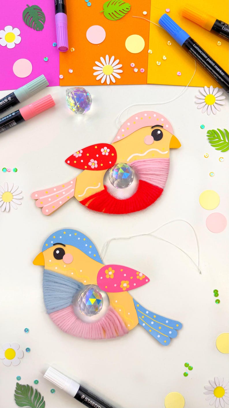 K&ouml;nnte beinhalten: Zwei handgefertigte Vogelornamente. Die V&ouml;gel haben gelbe K&ouml;rper, rote und rosa Akzente und blaue Fl&uuml;gel. Jeder Vogel hat einen kristallartigen Edelstein in der Mitte und ist mit rosa und blauer Wolle verziert. Der Hintergrund umfasst farbiges Papier und Bastelbedarf.