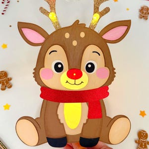 May include: A brown reindeer decoration with a red scarf and light-up antlers. The reindeer has a red nose and is surrounded by gingerbread men and star-shaped confetti. The antlers are adorned with small lights.