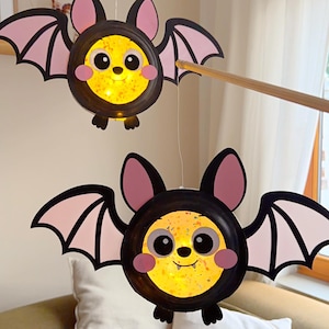 Könnte beinhalten: Zwei hängende Fledermaus-Dekorationen mit runden, beleuchteten gelben Körpern, schwarzen Rändern, großen Augen, rosa Wangen und kleinen Fangzähnen. Jede Fledermaus hat schwarz-rosa Flügel und ist an dünnen Schnüren an einem Holzstab befestigt.