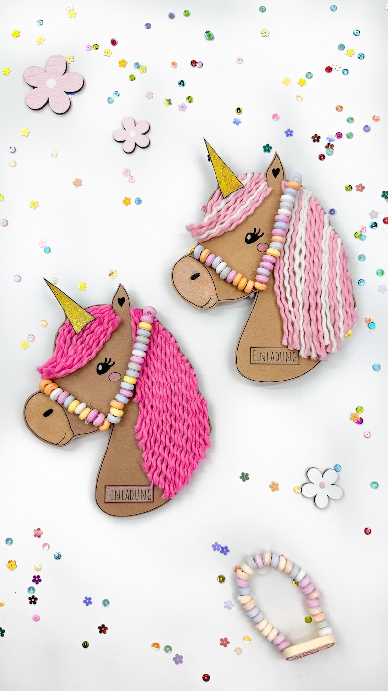Puede incluir: Dos manualidades de cabeza de unicornio de hilo rosa y blanco con collares de cuentas de caramelo y la palabra "Einladung" en la parte inferior. Las manualidades est&aacute;n sobre un fondo blanco con confeti de colores.