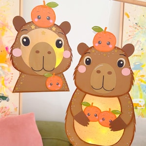 Può includere: Due decorazioni a sospensione a forma di capibara in materiale marrone con pance luminose. Ogni capibara presenta un volto da cartone animato con guance rosa e illustrazioni di piccoli frutti arancioni. Appese a bastoncini di legno.