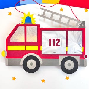 Puede incluir: Linterna de papel de camión de bomberos rojo con una franja amarilla, una escalera y el número 112 en el lateral. La linterna está rodeada de estrellas amarillas sobre un fondo blanco.