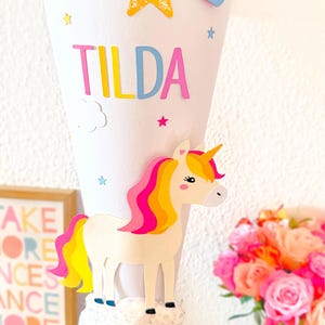 May include: A white cone with a blue base decorated with a unicorn, a rainbow, a star, and the name "TILDA".