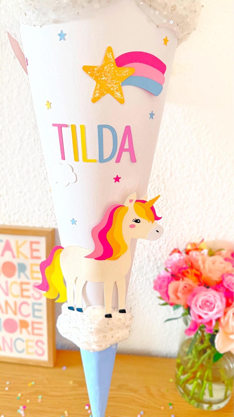May include: A white cone with a blue base decorated with a unicorn, a rainbow, a star, and the name "TILDA".
