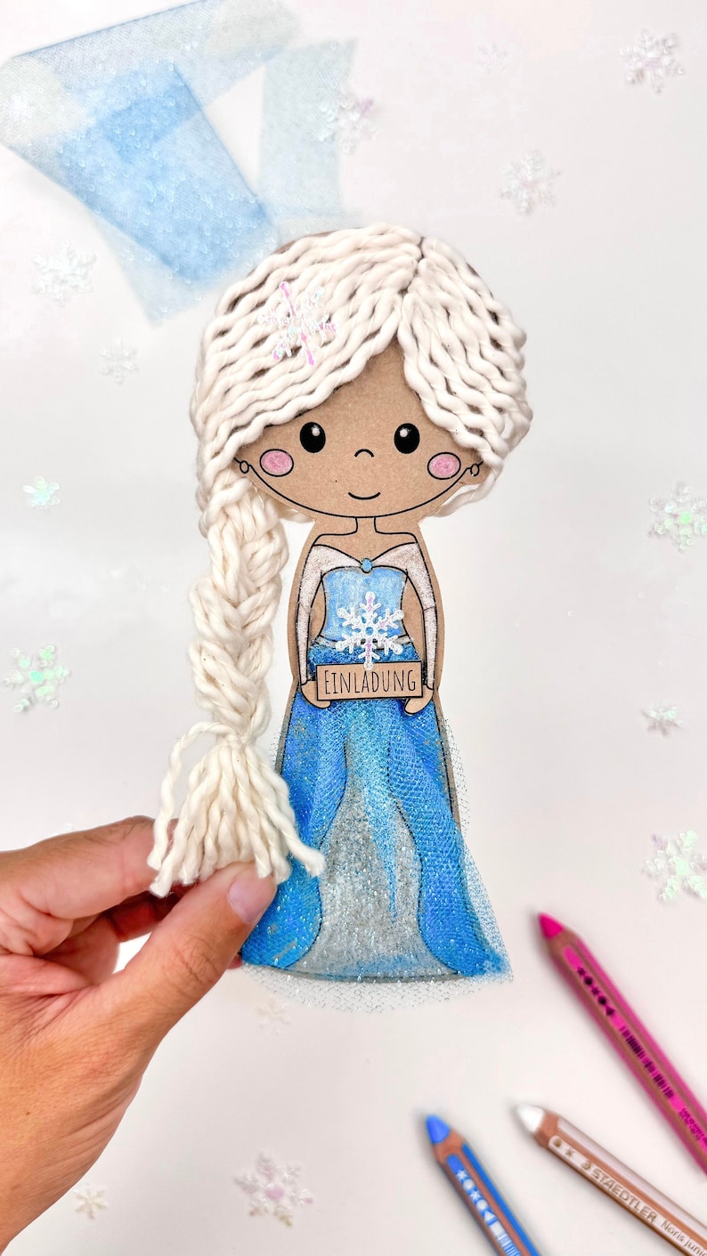 May include: A handmade paper doll of a girl with white yarn hair, wearing a blue and white dress with a snowflake design. She is holding a sign that says "Einladung" which is German for "Invitation".