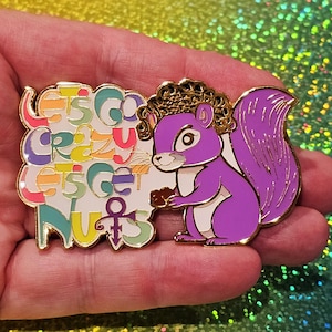 Puede incluir: Ardilla morada con una corona dorada sosteniendo una nuez marrón. La ardilla está sobre un fondo blanco con el texto "Let's Go Crazy Let's Get Nuts" en colores arcoíris y un símbolo morado.