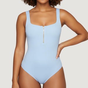 Puede incluir: Un bañador de una pieza azul claro con cuello cuadrado y tirantes anchos. El bañador tiene una cremallera dorada en la parte delantera. El bañador es de color liso. La modelo lleva el bañador.