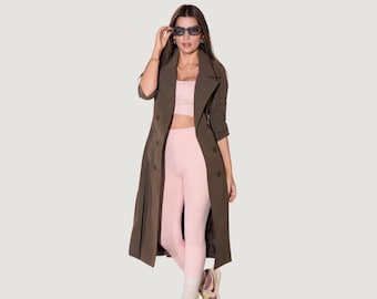 May include: A long olive green trench coat with a double-breasted button closure, worn over a light pink cropped top and matching high-waisted leggings. The outfit is paired with white socks and sneakers, and dark-tinted sunglasses are worn on the face.