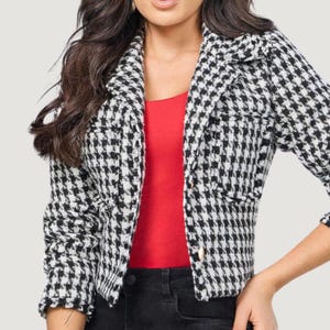 Può includere: Un blazer corto con motivo pied-de-poule bianco e nero, maniche lunghe, colletto con revers e chiusura frontale con bottoni. Il blazer è indossato sopra un top rosso senza maniche abbinato a pantaloni neri, creando un look a strati.