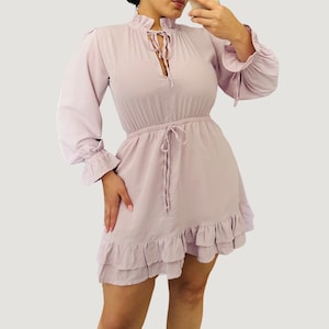 May include: A light purple dress with long sleeves and a ruffled hem. The dress features a high neckline with a tie detail and a drawstring waist. The sleeves have ruffled cuffs. The dress is made of a soft, flowing fabric.