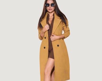 May include: A mustard yellow double-breasted long coat with a notched lapel and black buttons, worn over a dark brown zip-up top. The outfit is paired with dark-tinted sunglasses featuring a thin frame, creating a structured and layered look.
