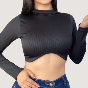 May include: Black long-sleeve crop top with a mock neck. The top has a ribbed texture on the front and smooth fabric on the sleeves. The bottom hem has a unique curved design. The model is wearing blue jeans.