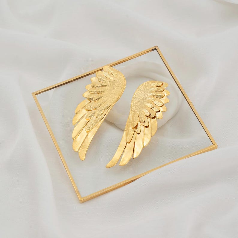 Elegant angel wing earrings made of bronze with gold plating. Drop design with curved wing shape, engraved feather texture, and glossy polished finish. Bright gold tone creates a timeless and graceful statement look.