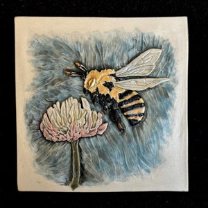 May include: A ceramic tile featuring a detailed relief sculpture of a bee pollinating a clover flower. The bee is yellow and black with gold accents, and the flower is white with pink accents. The background is a textured blue.