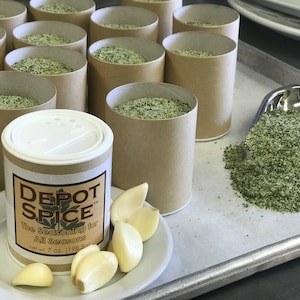 May include: A brown cardboard container with a white lid filled with green seasoning. The container is labeled "Depot Spice" and "The Seasoning for All Seasons". The container is sitting on a white surface with a small plate of garlic cloves.