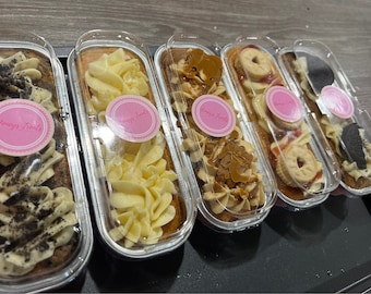 May include: Several rectangular cakes in clear plastic containers. Each cake is topped with different frostings and toppings, including cookies, caramel, and sprinkles. Pink stickers with the text "Honey's Treats" are on each container.