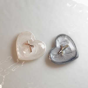 Puede incluir: Dos platos en forma de corazón, uno blanco y otro plateado, cada uno con una cruz plateada. El plato blanco tiene un brillo nacarado, mientras que el plato plateado tiene un brillo metálico. Ideales para pequeñas joyas o recuerdos.