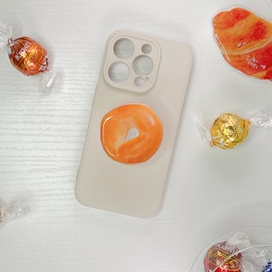 Könnte beinhalten: Eine weiße Handyhülle mit einem donutförmigen Handygriff. Der Griff ist orange und hat eine weiße Mitte. Die Handyhülle hat einen Kameraobjektiv-Ausschnitt mit drei Kreisen. Im Hintergrund befinden sich mehrere Schokoladenbonbons.