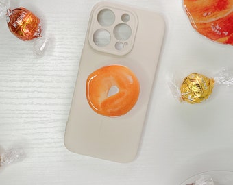May include: A white phone case with a donut-shaped phone grip. The grip is orange and has a white center. The phone case has a camera lens cutout with three circles. There are several chocolate candies in the background.