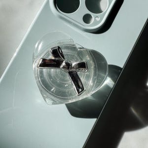 May include: A clear, heart-shaped phone grip with a silver bow design, attached to a light blue phone. The phone has a triple-lens camera system. The grip is transparent, allowing the phone's colour to show through.