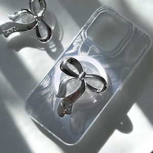 May include: A clear phone case with a silver bow ring holder. The case has a textured, brushed finish. The bow ring holder is attached to the back of the case.