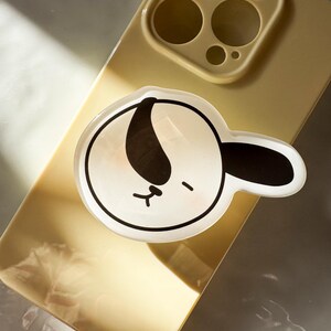 May include: A clear, circular phone grip featuring a cartoon dog face with black and white accents. The dog has a closed-eye expression and a pink blush. The grip is attached to a gold phone case.