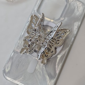 May include: A clear phone case with a silver butterfly phone ring holder. The case has a shimmery, iridescent finish.