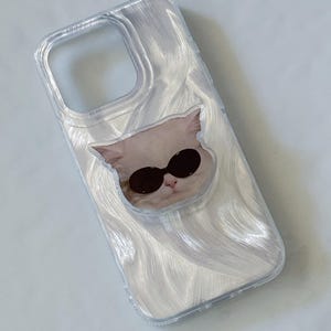 May include: A clear phone case with a light-colored, wavy design. The case features a graphic of a cat wearing black sunglasses. The camera cutout is visible at the top.