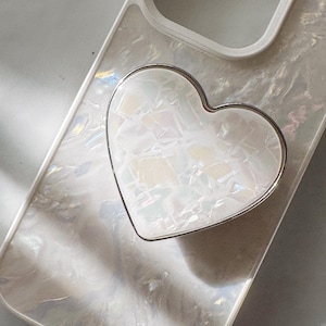 May include: A white iridescent phone case with a silver heart-shaped phone grip.