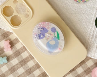 May include: A cream-coloured phone case with a clear phone grip featuring a floral design with purple flowers, a blue bird, and white pearl-like beads.