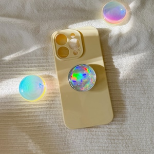 Puede incluir: Una funda de teléfono beige con un pop socket. El pop socket es iridiscente y tiene un efecto arcoíris. La funda del teléfono tiene un recorte para el objetivo de la cámara.