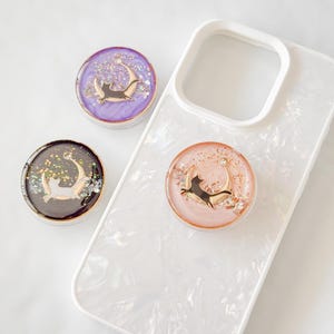 Può includere: Tre supporti per telefono con un design a forma di luna crescente e gatto. I supporti sono realizzati in resina trasparente con glitter e sono disponibili in viola, nero e rosa.