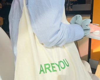May include: A white canvas tote bag with a green "ARE YOU" graphic printed on the front. The bag is being worn by a person wearing a blue and white striped shirt.